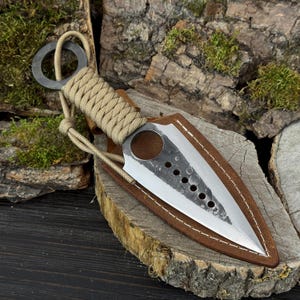 Op de afbeelding: Een tweesnijdend metalen lemmet met een puntige top en een reeks cirkelvormige uitsparingen in het midden. Het handvat is omwikkeld met beige paracord en heeft een ring aan het uiteinde. Het lemmet zit gedeeltelijk in een bruine lederen schede.
