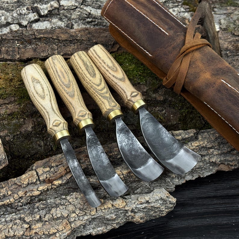 May include: Four wood carving chisels with light wood handles and metal blades are displayed on a textured wooden surface. The chisels have brass collars and are accompanied by a rolled brown leather tool roll.