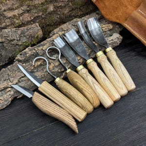 Pode incluir: Um conjunto de ferramentas para entalhar madeira com cabos de madeira e lâminas de metal. As lâminas têm várias formas, incluindo goivas curvas e facas retas. As ferramentas estão dispostas sobre uma superfície de madeira escura.