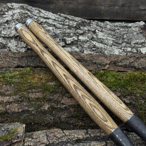 Może przedstawiać: Dwa narzędzia z drewnianymi uchwytami i metalowymi końc&oacute;wkami. Uchwyty mają jasnobrązowy wz&oacute;r słoj&oacute;w drewna. Metalowe końc&oacute;wki są ciemnoszare. Narzędzia są ustawione pod kątem na teksturowanej powierzchni przypominającej korę z plamami zielonego mchu.