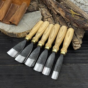 May include: A set of six wood carving chisels with light wood handles and silver blades. The chisels are arranged in a row, with a leather tool roll in the background. The blades have a dark, textured appearance.