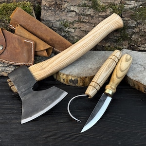 May include: Axe and two knives with wooden handles and metal blades. The axe has a dark metal head and a light-colored wooden handle. The knives have light-colored wooden handles and metal blades. Leather sheaths are also included.