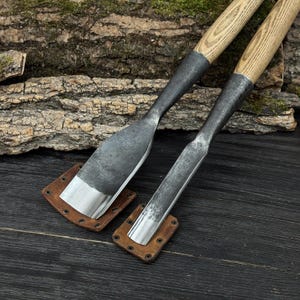 Może przedstawiać: Dwa metalowe dłuta z drewnianymi uchwytami i sk&oacute;rzanymi pochwami. Dłuta mają srebrne ostrze i ciemny metalowy korpus. Drewniane uchwyty są jasnobrązowe. Sk&oacute;rzane pochwy są brązowe i kwadratowe. Tło to ciemna drewniana powierzchnia.
