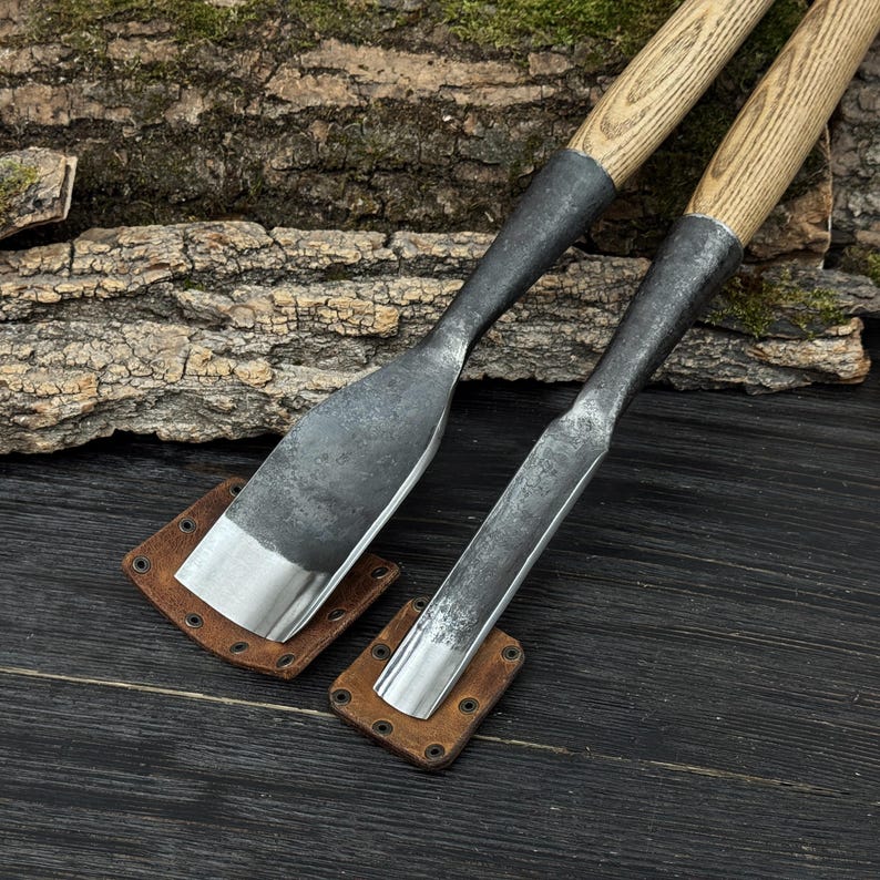 Może przedstawiać: Dwa metalowe dłuta z drewnianymi uchwytami i sk&oacute;rzanymi pochwami. Dłuta mają srebrne ostrze i ciemny metalowy korpus. Drewniane uchwyty są jasnobrązowe. Sk&oacute;rzane pochwy są brązowe i kwadratowe. Tło to ciemna drewniana powierzchnia.