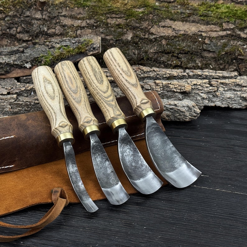 May include: Set of four wood carving gouges with light wood handles, brass ferrules, and curved steel blades. The tools are arranged on a brown leather sheath, with a natural wood background.