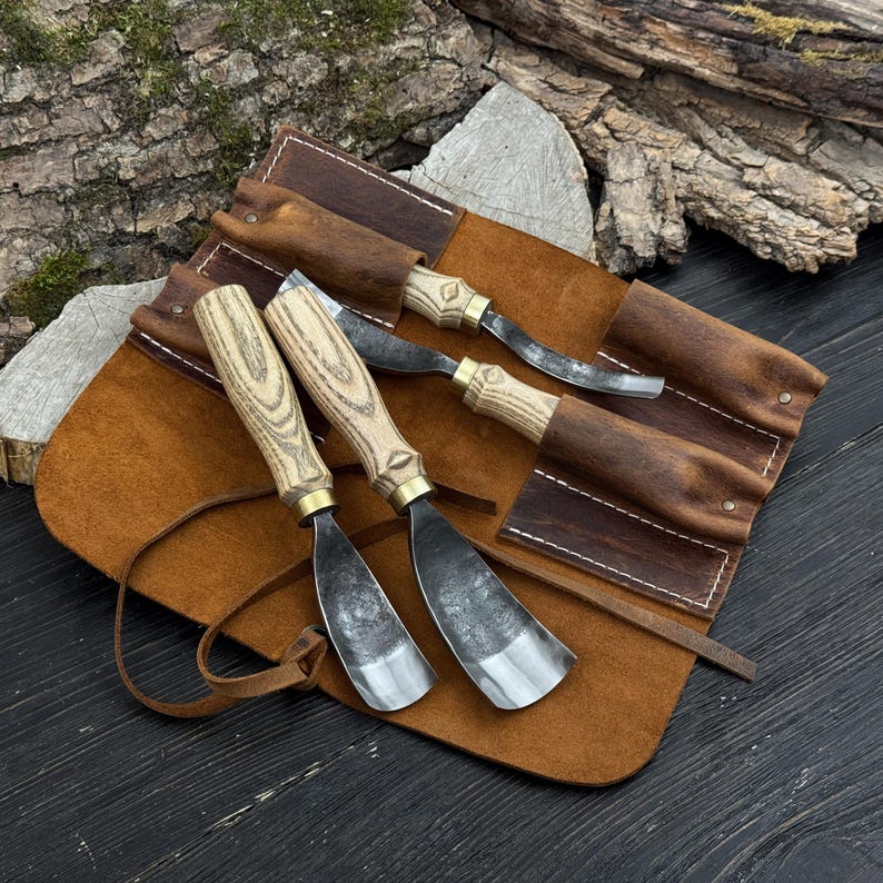 May include: A collection of wood carving tools with wooden handles and metal blades, presented in a brown leather tool roll. The tools feature a natural wood grain finish and brass details. The leather roll is secured with a leather strap.