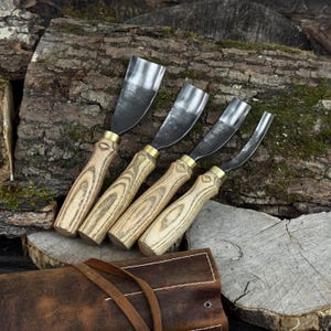 May include: A set of four wood carving gouges with curved blades. The gouges have light-coloured wooden handles with brass ferrules. The blades are dark metal. A brown leather tool roll is partially visible. The tools are arranged on a wood surface.