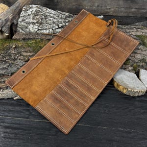 May include: A brown leather tool roll with multiple pockets and white stitching. The roll is open, revealing a suede interior and a leather tie closure. The leather has a distressed appearance.