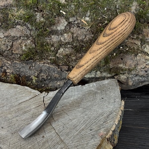 May include: A woodworking gouge with a curved, silver-colored blade and a light brown wooden handle. The handle has a rounded end and is made of wood with a visible grain pattern. The tool is designed for carving and shaping wood.