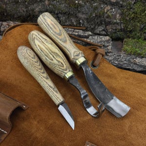 May include: Three woodworking tools with light-coloured, textured wooden handles and brass ferrules. The set includes a small pointed carving knife, a curved hook knife, and a wider gouge, all featuring dark, forged metal blades.
