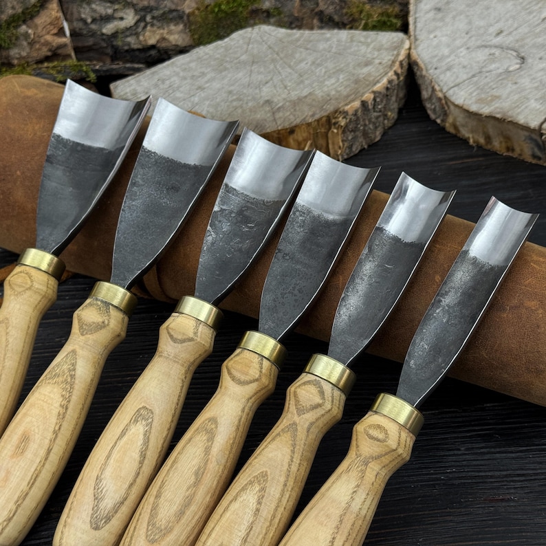 May include: A set of six wood carving gouges with curved, steel blades. The handles are light-colored wood with brass ferrules. The blades have a dark, textured appearance.