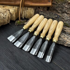 May include: Set of six wood carving chisels with light wood handles, brass ferrules, and silver blades. The tools are arranged in a row on a dark wooden surface, with a leather tool roll in the background.