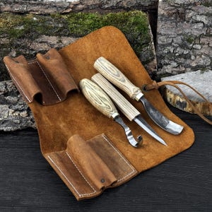 Wood carving kit with leather roll, hook knife, sloyd knife and curved gouge displayed together.