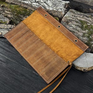 May include: A brown leather tool roll with multiple pockets and white stitching. The roll is open, revealing a tan interior. Leather ties secure the roll. The background includes wood.