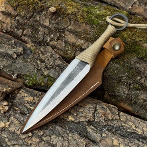 Può includere: Un pugnale in acciaio a doppio taglio con una finitura grigio scuro testurizzata al centro e un bordo lucido. L'impugnatura è avvolta in paracord color marrone chiaro con un anello metallico all'estremità. È abbinato a un fodero in pelle marrone con bottone a pressione.