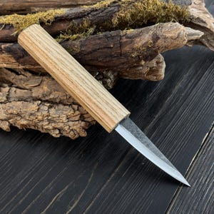 Sloyd carving knife with ash wood handle and narrow carbon steel blade, photographed on bark and moss.
