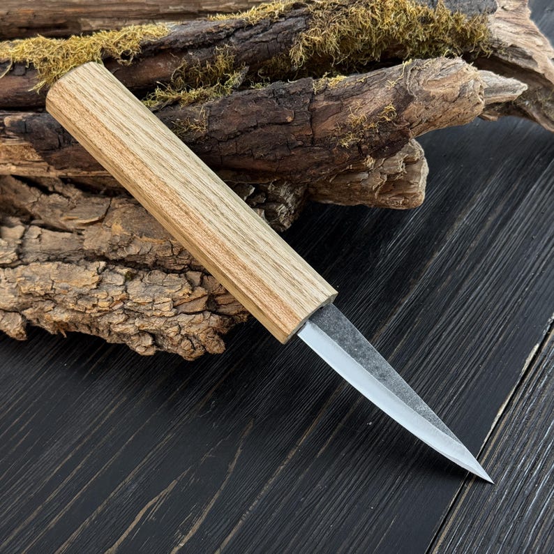 Sloyd carving knife with ash wood handle and narrow carbon steel blade, photographed on bark and moss.