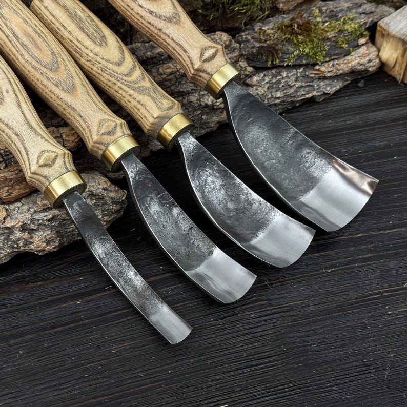May include: Four wood carving gouges with wooden handles and metal blades. The handles are light brown with a brass ferrule. The curved blades have a silver finish. The tools are displayed on a dark wooden surface.