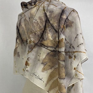 May include: A white silk scarf with a botanical print in shades of brown, beige, and gray. The scarf is folded and draped over a mannequin. The artist's signature, "Laura Baker", is visible in the lower right corner.