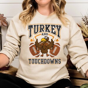 Könnte beinhalten: Ein beiger Pullover mit einem Grafikdruck eines Truthahns, der zwei Fußbälle hält. Der Text "TURKEY AND TOUCHDOWNS" ist unter dem Truthahn gedruckt.