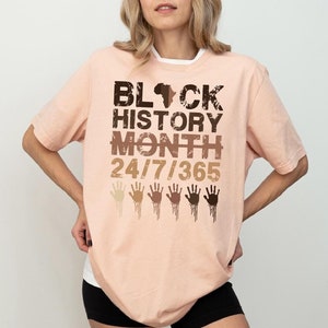 May include: A peach-colored t-shirt with the text "Black History Month 24/7/365" in black and brown lettering. The shirt also features a silhouette of the continent of Africa and a row of five handprints in various shades of brown.