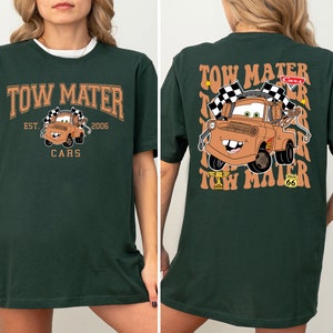 K&ouml;nnte beinhalten: Gr&uuml;nes T-Shirt mit einem braunen Cartoon-Abschleppwagen auf der Vorderseite und dem Text "Tow Mater Est. 2006 Cars". Die R&uuml;ckseite des Shirts hat den Text "Tow Mater" in einem wellenf&ouml;rmigen Muster wiederholt, mit einem braunen Cartoon-Abschleppwagen in der Mitte.