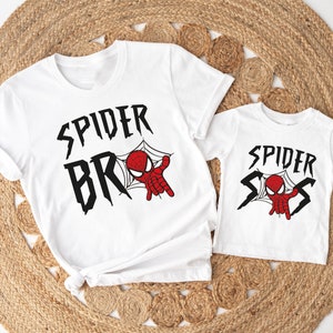 May include: Two white t-shirts with a Spider-Man design. The larger shirt says "Spider Bros" with a cartoon Spider-Man in a web. The smaller shirt says "Spider Sis" with a cartoon Spider-Man in a web.