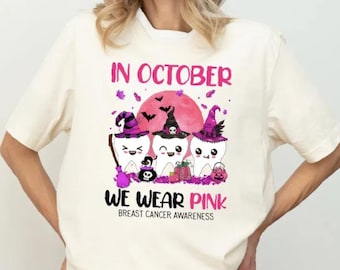 Puede incluir: Camiseta color crema con un diseño gráfico. El diseño presenta tres dientes de dibujos animados con sombreros de bruja, una luna rosa y el texto "IN OCTOBER WE WEAR PINK BREAST CANCER AWARENESS".