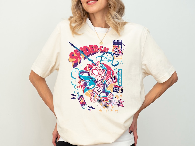 K&ouml;nnte beinhalten: Ein wei&szlig;es T-Shirt mit einem bunten Grafikdruck eines Katers, der ein Spiderman-Kost&uuml;m tr&auml;gt. Die Grafik enth&auml;lt den Text "SPIDER-CAT" und "FREE STICKERS".