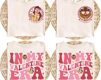 以下が含まれることがあります： バレンタインデーのデザインが施された、クリーム色のTシャツ4枚。2枚のシャツには、ハート型のメガネをかけたベルと野獣が描かれ、「I'M HIS BEAUTY」と「I'M HER BEAST」というフレーズが添えられています。他の2枚のシャツには「IN MY VALENTINE ERA」と書かれています。