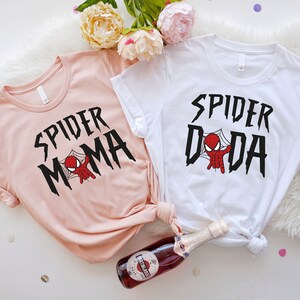 May include: Two t-shirts with a pink and white color scheme. The pink shirt has the text "SPIDER MAMA" with a cartoon spider-man in a web. The white shirt has the text "SPIDER DADA" with a cartoon spider-man in a web.