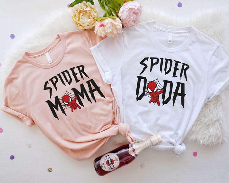 May include: Two t-shirts with a pink and white color scheme. The pink shirt has the text "SPIDER MAMA" with a cartoon spider-man in a web. The white shirt has the text "SPIDER DADA" with a cartoon spider-man in a web.