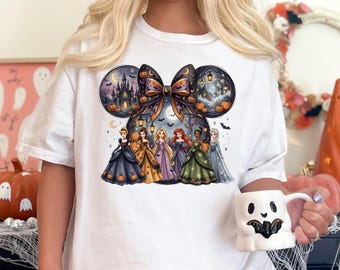 Op de afbeelding: Een wit t-shirt met korte mouwen en een afbeelding van zes geanimeerde prinsessen in baljurken onder een grote strik en een Mickey Mouse-vormige omtrek met een spookkasteel, pompoenen, lantaarns en vleermuizen tegen een nachtelijke hemel.