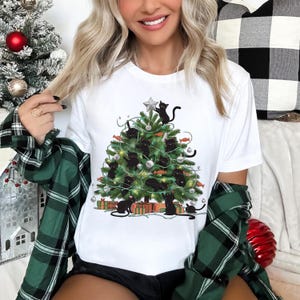 Pode incluir: Uma t-shirt branca de manga curta com uma estampa gráfica de uma árvore de Natal verde decorada com ornamentos prateados, luzes e vários gatos pretos a subir pelos ramos, rematada com uma estrela prateada e caixas de presente.