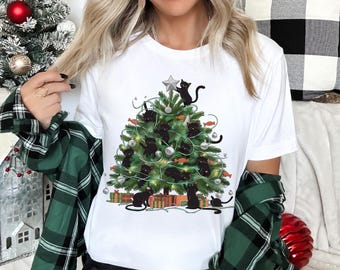 Op de afbeelding: Een wit t-shirt met korte mouwen en een grafische print van een groene kerstboom versierd met zilveren ornamenten, kerstverlichting en meerdere zwarte katten die in de takken klimmen, bekroond met een zilveren ster en cadeauverpakkingen.