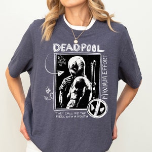 May include: A dark blue t-shirt with a white graphic of Deadpool, a comic book character. The graphic includes the text "Deadpool Maximum Effort" and "They call me the merc with a mouth".