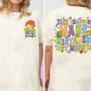Op de afbeelding: Een off-white t-shirt met een kleine afbeelding op de linkerborst van een roodharig personage met een groene hagedis en de naam Jennifer. De achterkant toont de kleurrijke tekst Take Chances Make Mistakes Get Messy met diverse schooliconen.
