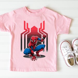 May include: Pink t-shirt with a cartoon image of Spiderman in a crouching pose. The image is set against a red and black web design.