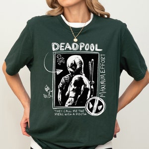 May include: A dark green t-shirt with a white graphic print of Deadpool. The graphic features Deadpool in a black and white illustration with the text "Deadpool Maximum Effort They Call Me The Merc With A Mouth".