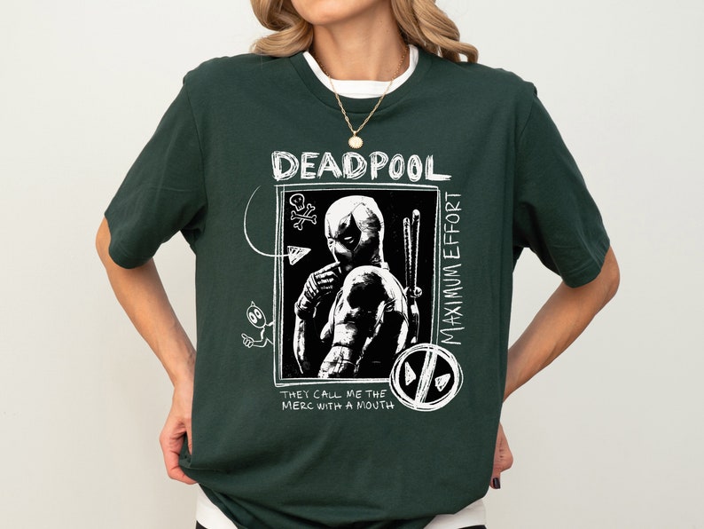May include: A dark green t-shirt with a white graphic print of Deadpool. The graphic features Deadpool in a black and white illustration with the text "Deadpool Maximum Effort They Call Me The Merc With A Mouth".