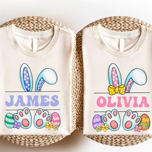 May include: Two off-white t-shirts featuring a graphic of blue bunny ears and feet. The left shirt displays the name JAMES in purple, while the right shirt displays the name OLIVIA in pink with a yellow bow. Both include colorful Easter eggs.
