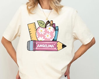 May include: A cream-colored t-shirt featuring a graphic print of a pink apple with white daisy patterns, a yellow ruler, and colorful crayons. A large pink pencil is positioned horizontally below the apple with the name ANGELINA printed in white.