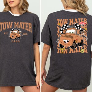 K&ouml;nnte beinhalten: Ein dunkelgraues T-Shirt mit einem Cartoon-Bild von Tow Mater aus dem Disney Pixar Film Cars. Die Vorderseite des Shirts zeigt "Tow Mater" mit "Est. 2006" und "Cars" darunter. Die R&uuml;ckseite des Shirts zeigt ein gr&ouml;&szlig;eres Bild von Tow Mater mit dem Text "Tow Mater" in einer Retro-Schrift.
