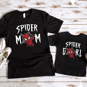May include: Two black t-shirts with a spider-man graphic. The larger shirt says "Spider Mom" and the smaller shirt says "Spider Girl".