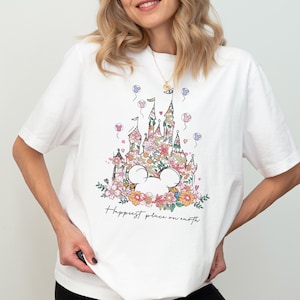 Pu&ograve; includere: Maglietta bianca con una stampa floreale del castello di Cenerentola e il testo "Happiest place on earth".