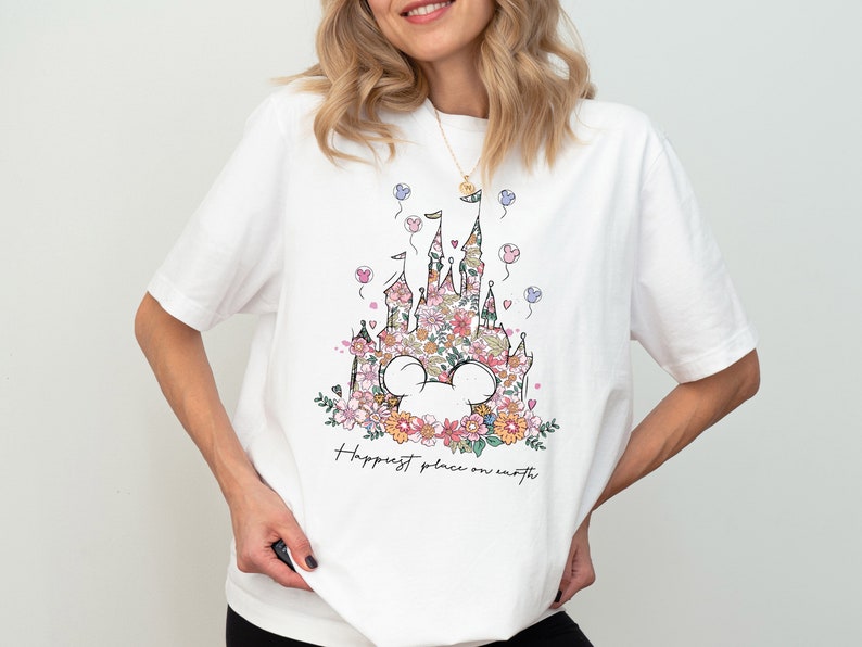 Pu&ograve; includere: Maglietta bianca con una stampa floreale del castello di Cenerentola e il testo "Happiest place on earth".