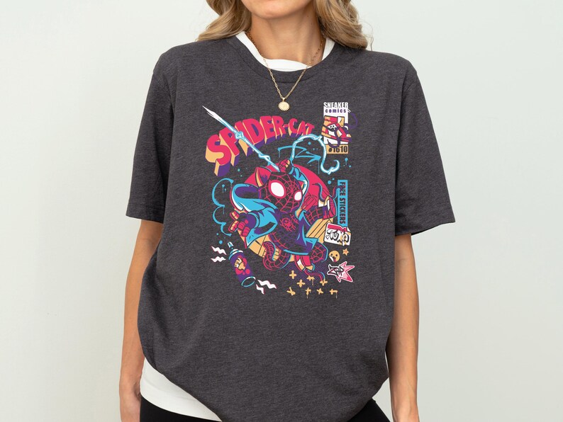 K&ouml;nnte beinhalten: Ein dunkelgraues T-Shirt mit einem Grafikdruck eines Comic-Spider-Man-Charakters in einem rot-schwarzen Anzug. Die Grafik ist von buntem Comic-Text und -Bildern umgeben. Der Text auf dem Shirt lautet "Spider-Cat".