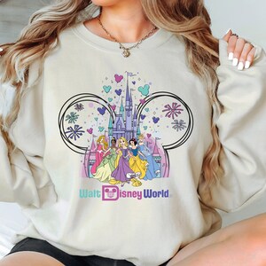 K&ouml;nnte beinhalten: Ein hellbeiger Pullover mit einem grafischen Druck von Aschenputtel, Schneewittchen, Arielle, Belle und Tiana, die vor dem Schloss von Aschenputtel stehen. Die Grafik ist von Mickey-Maus-Ohren und Feuerwerk umgeben. Der Text "Walt Disney World" ist unter der Grafik gedruckt.