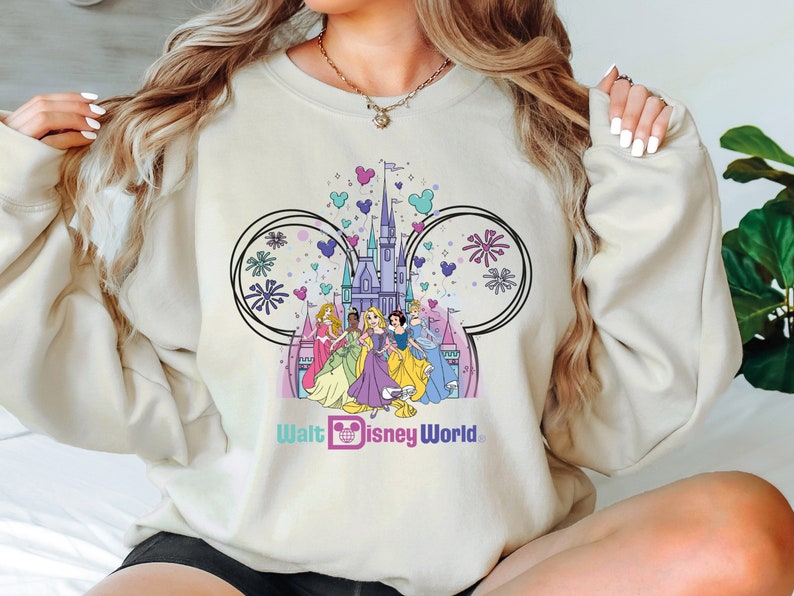 K&ouml;nnte beinhalten: Ein hellbeiger Pullover mit einem grafischen Druck von Aschenputtel, Schneewittchen, Arielle, Belle und Tiana, die vor dem Schloss von Aschenputtel stehen. Die Grafik ist von Mickey-Maus-Ohren und Feuerwerk umgeben. Der Text "Walt Disney World" ist unter der Grafik gedruckt.