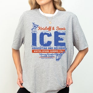 K&ouml;nnte beinhalten: Graues T-Shirt mit einer blauen und roten Grafik von Olaf und Sven, die Eis liefern. Der Text lautet "Kristoff & Sven's ICE HARVESTING AND DELIVERY WINTER; SPRING SUMMER FALL! Delivering throughout Arendelle Kingdom."